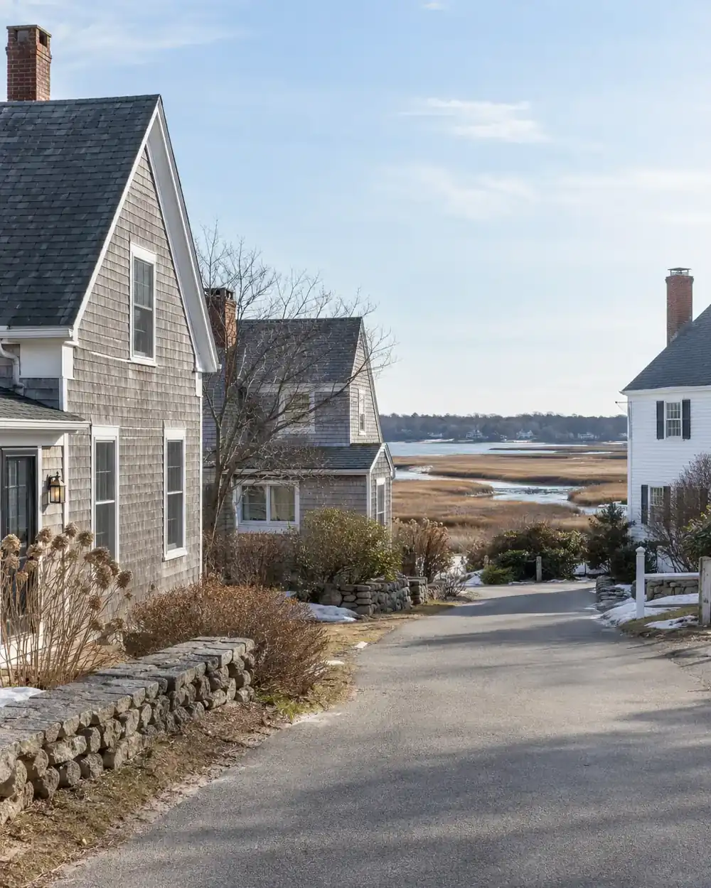 Shingled cottages and a white clapboard house beside a quiet road and salt marsh.