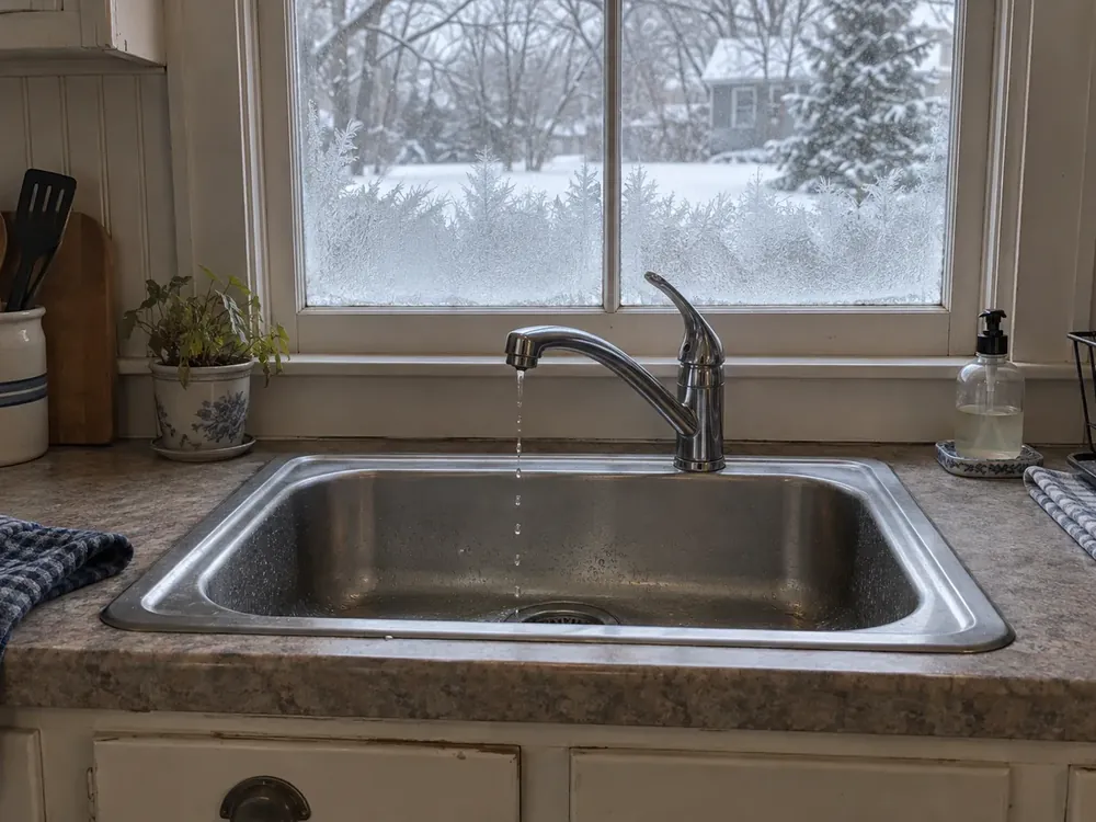 Kitchen faucet turned fully on gives only a thin trickle into the sink beneath a frosted winter window.